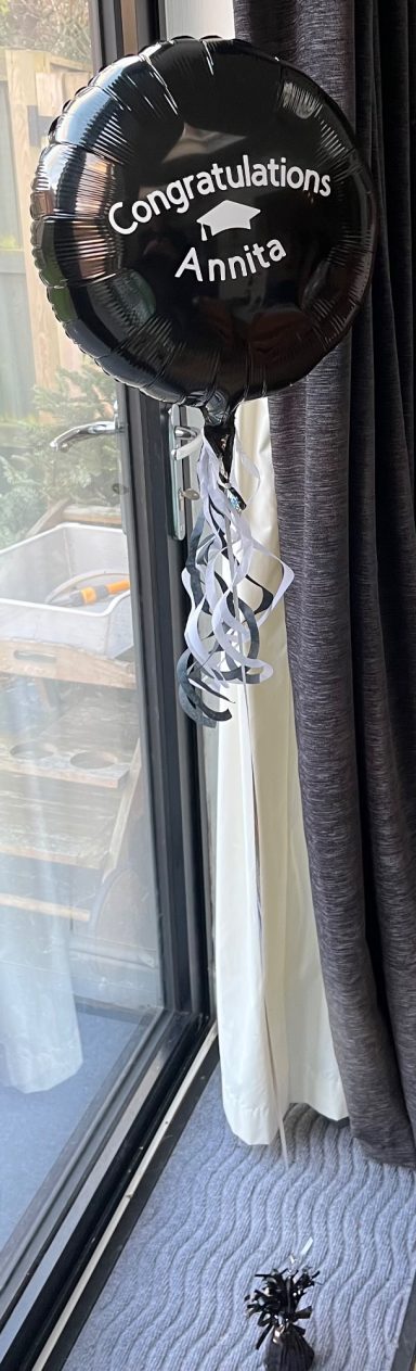 Black balloon with "Congratulations" text tied with silver ribbons, near a window.