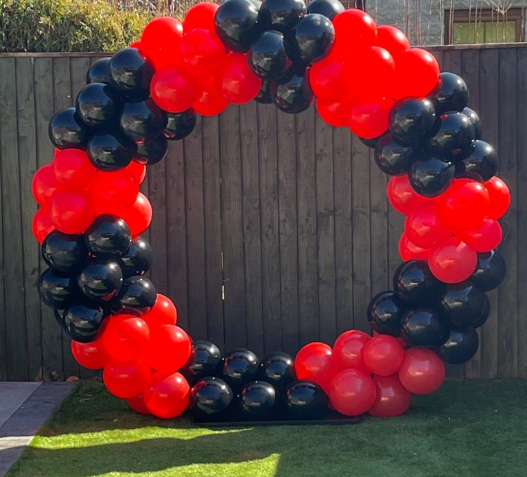 Red and black balloon arch set against a wooden fence in a garden.