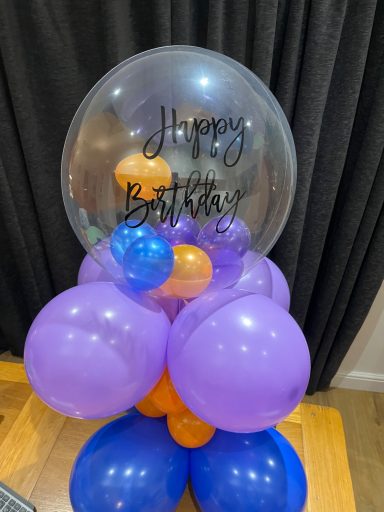 A balloon arrangement in purple, orange, and blue with a clear balloon saying "Happy Birthday."