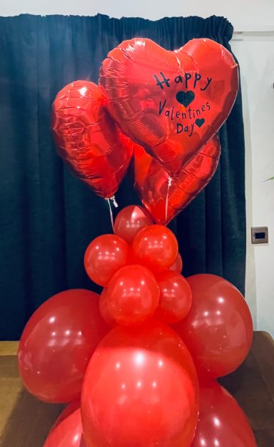 A cluster of red heart-shaped balloons with "Happy" written on them, surrounded by round balloons.